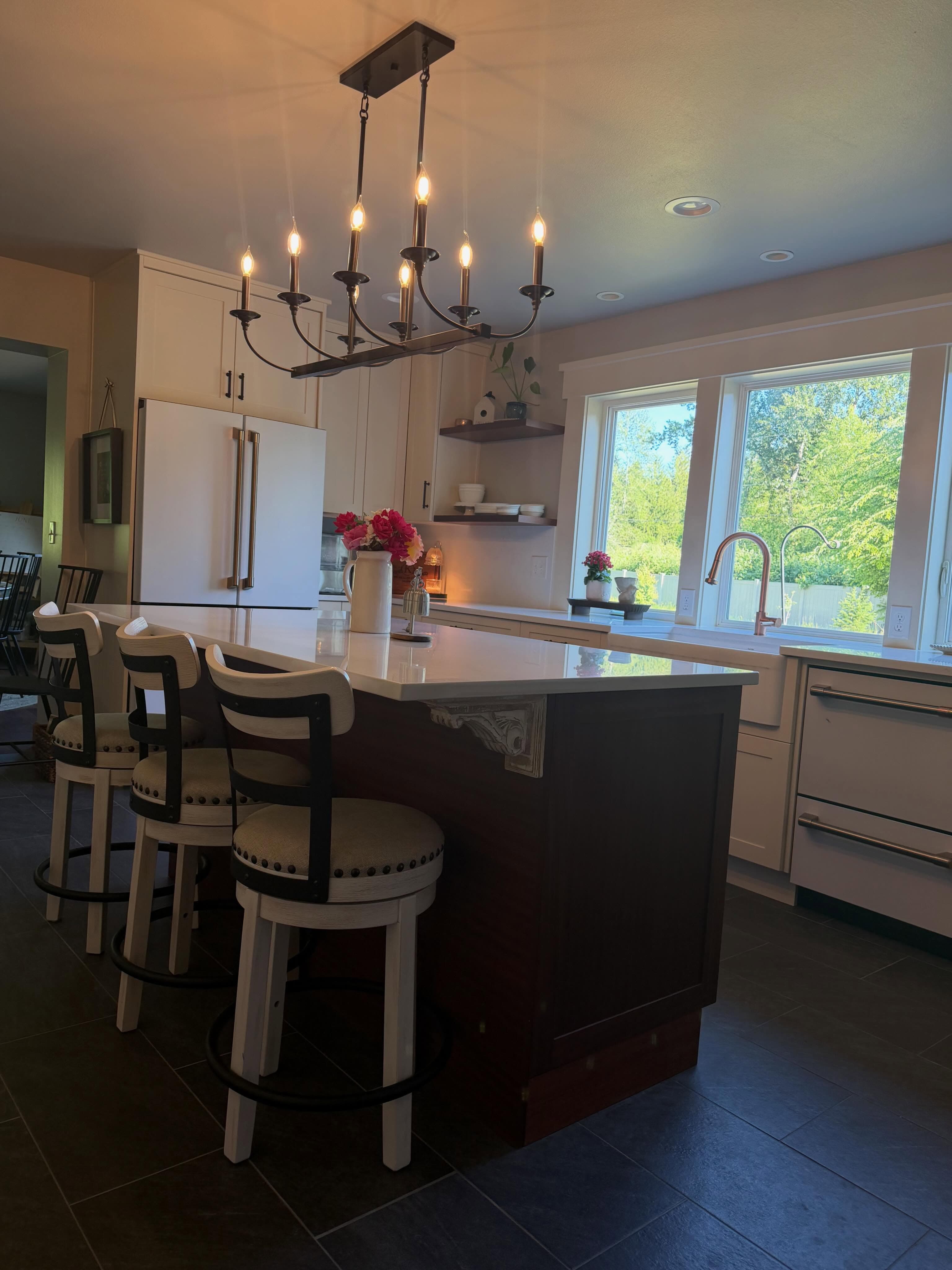 A finished white-shaker kitchen remodel with a dark walnut island, quartz countertop, oil-rubbed-bronze faucet and candelabra pendant, PNW greenery through the windows.