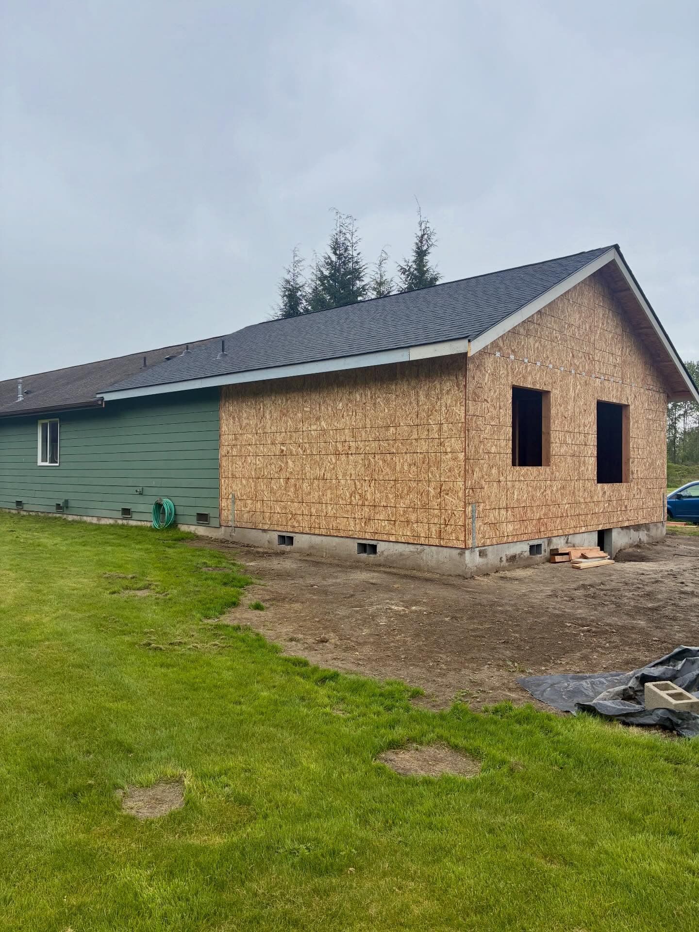 A home addition in progress — raw OSB sheathing on the new structure beside the existing sage-green siding, fresh concrete foundation, green lawn.