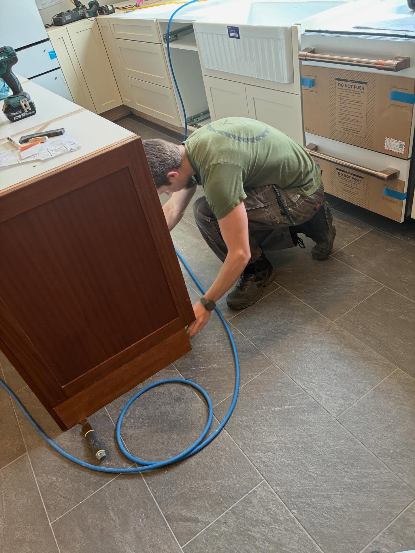 A Carmichael Carpentry crew member kneeling to set a walnut cabinet panel in a white-shaker kitchen, slate-grey tile underfoot.