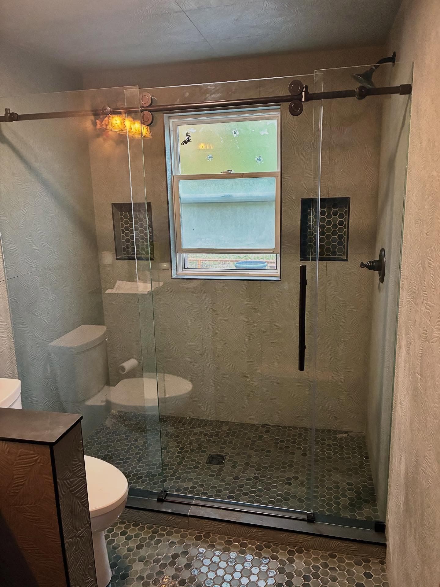 A finished bathroom remodel with sage-celadon tile, oil-rubbed-bronze barn-door shower hardware, a grey-green hex floor and an amber sconce.
